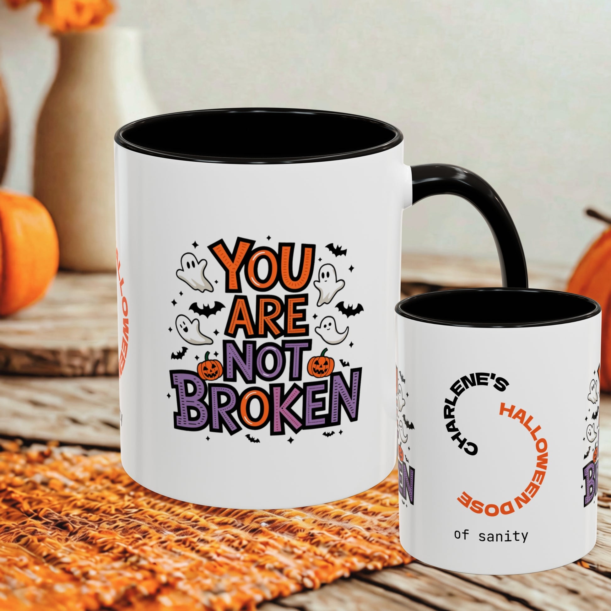 A white ceramic mug with a black interior and handle is shown alongside a smaller matching mug. The larger mug features a Halloween-themed graphic reading 'YOU ARE NOT BROKEN' with ghosts, bats, pumpkins, and spiders.