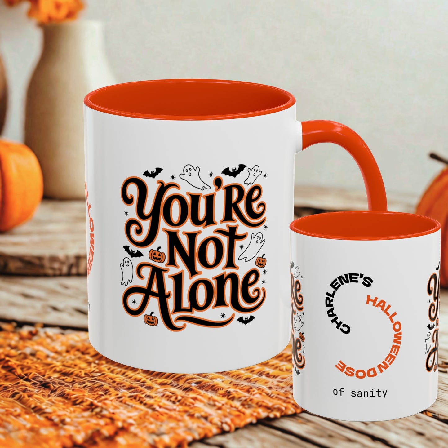 A white ceramic mug with a bright orange interior and handle, printed with the phrase 'You're Not Alone' in stylized orange and black lettering with small Halloween motifs. A smaller matching mug is shown beside it on a rustic table with autumn decor in the background.