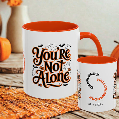 A white ceramic mug with a bright orange interior and handle, printed with the phrase 'You're Not Alone' in stylized orange and black lettering with small Halloween motifs. A smaller matching mug is shown beside it on a rustic table with autumn decor in the background.