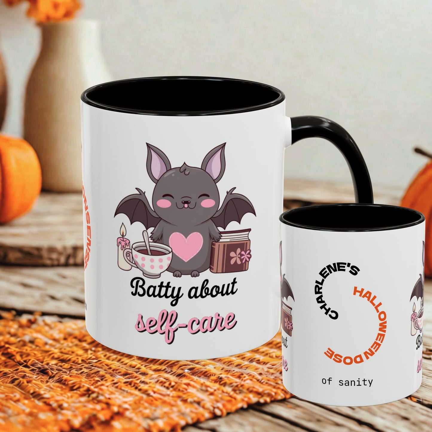Two white ceramic mugs with black interiors and handles, shown on a rustic wooden surface with autumn props. The larger mug features a cute bat graphic with a pink heart and the text 'Batty about self-care.