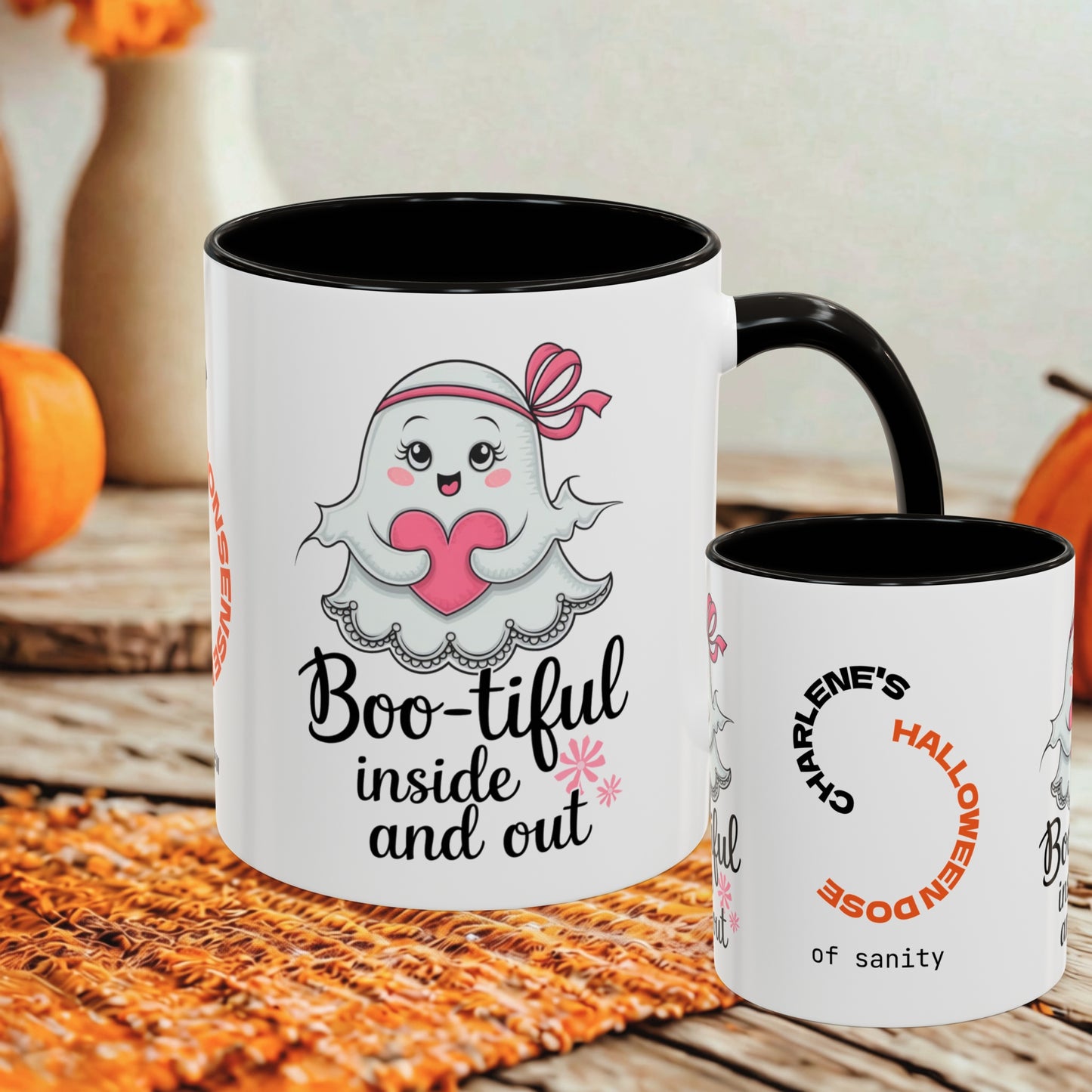 Two white ceramic mugs with black interiors and handles, displayed on a rustic wooden surface with autumn props. The larger mug features a cute cartoon ghost holding a pink heart with the text 'Boo-tiful inside and out', and the smaller mug reads 'Charlene's Halloween dose of sanity'.