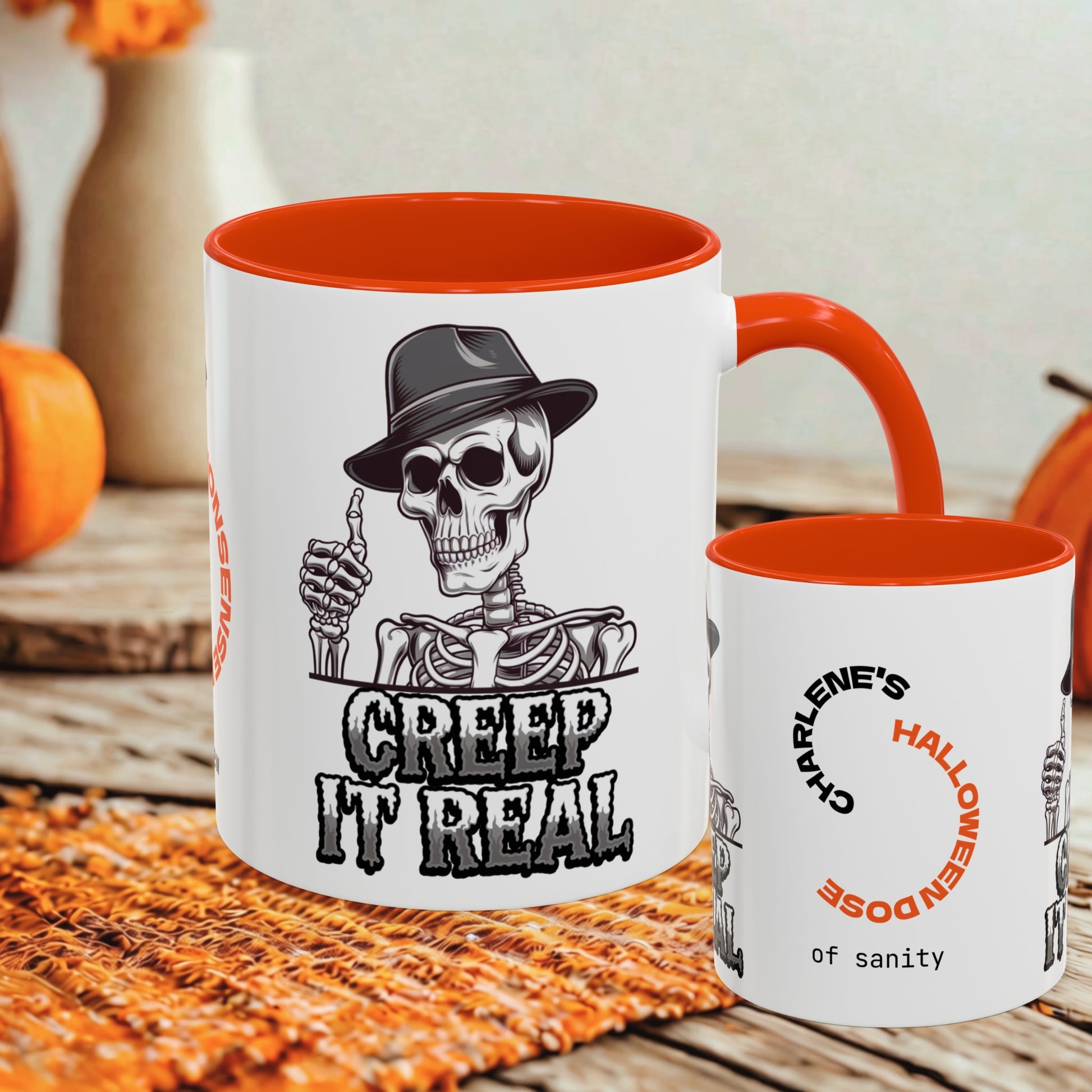 A white ceramic coffee mug with a bright orange interior and handle, featuring a black-and-white skeleton graphic wearing a hat and the text 'CREEP IT REAL.' A smaller matching mug stands beside it on a rustic wooden surface with autumn decor in the background.
