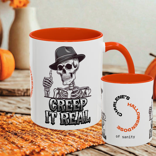 A white ceramic coffee mug with a bright orange interior and handle, featuring a black-and-white skeleton graphic wearing a hat and the text 'CREEP IT REAL.' A smaller matching mug stands beside it on a rustic wooden surface with autumn decor in the background.