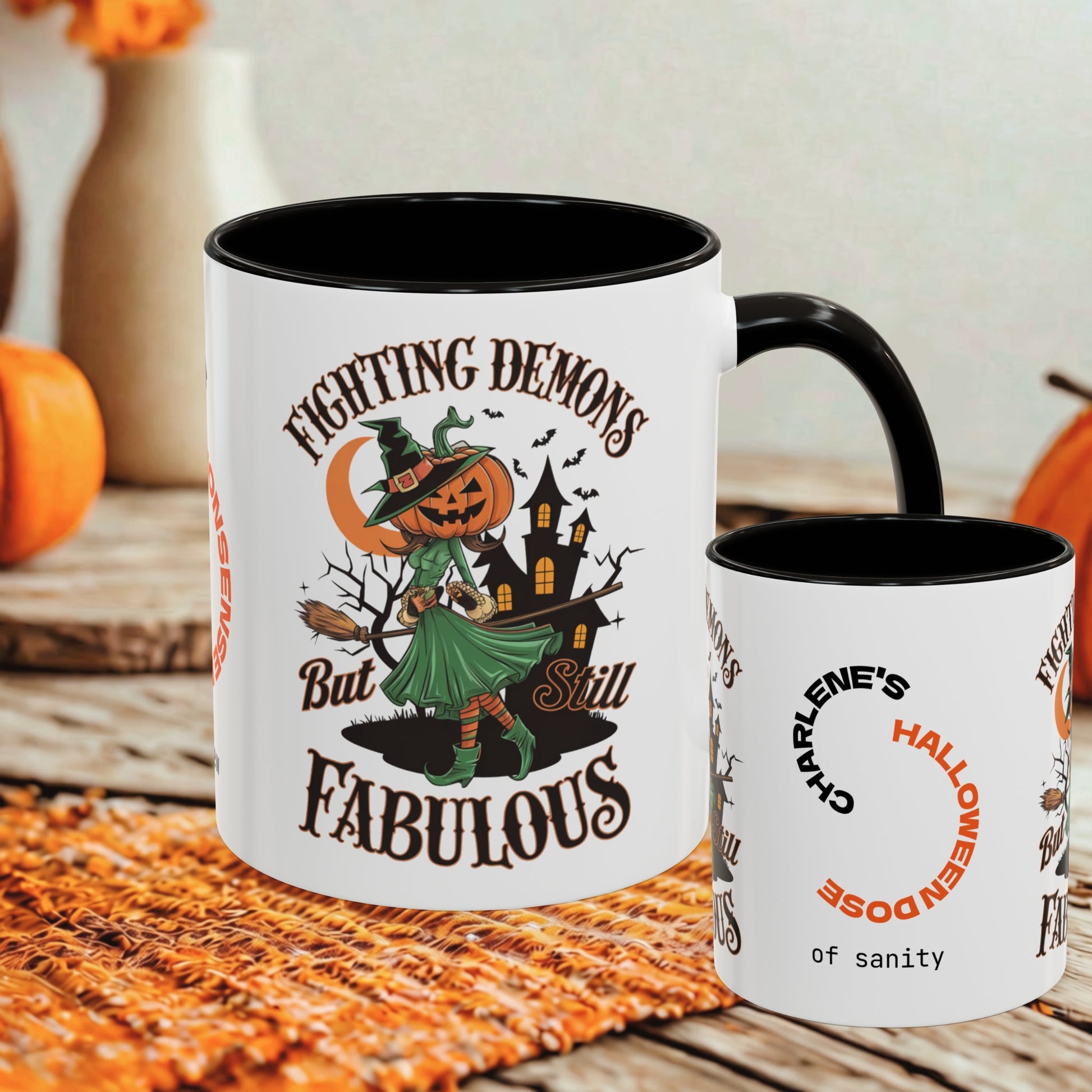 A white ceramic Halloween-themed mug with a black handle and interior, shown alongside a smaller side view of the same mug. The front design features a witch in a green dress with a pumpkin head, a haunted house, bats, and the text 'FIGHTING DEMONS But Still FABULOUS.