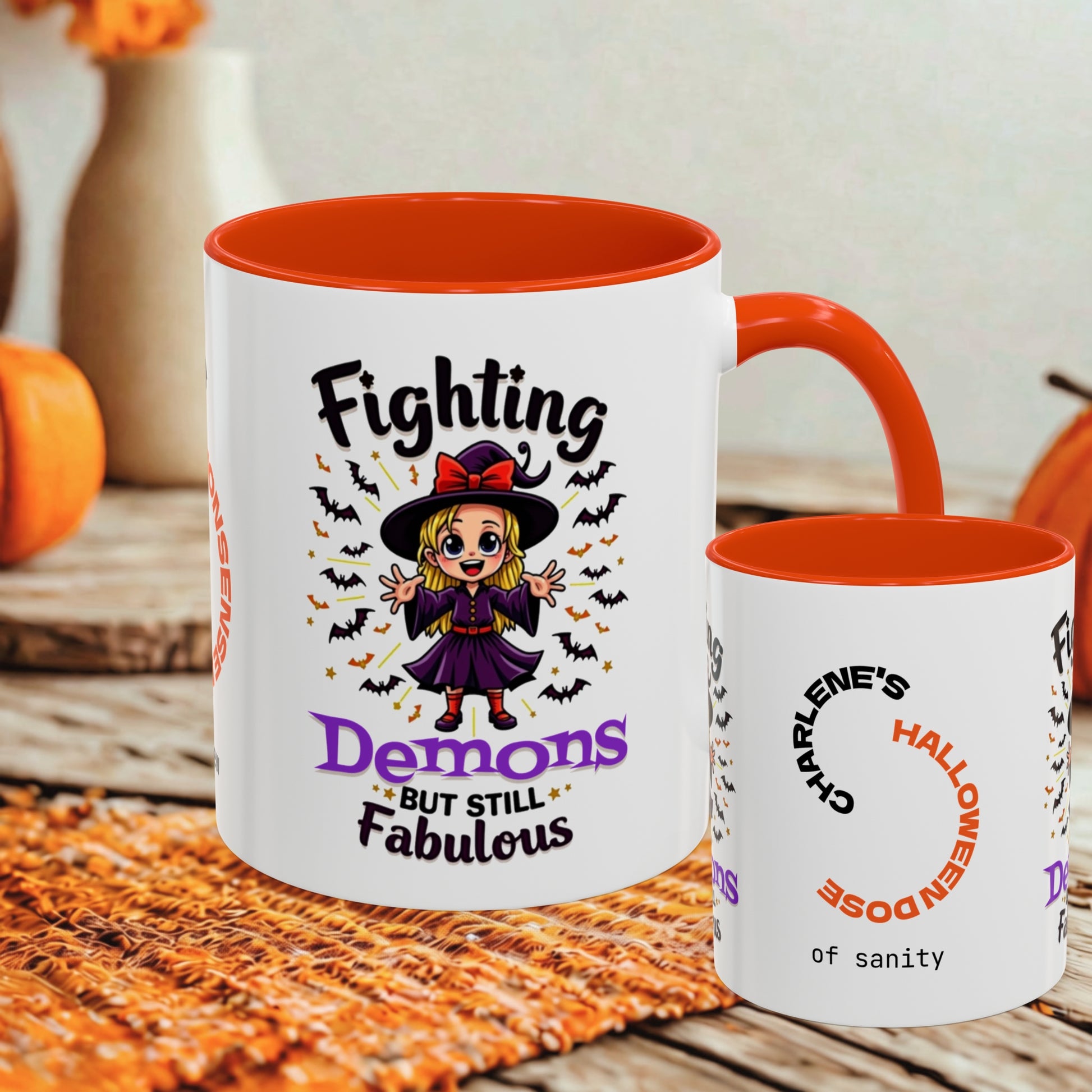 Two white ceramic mugs with orange interiors and handles are shown on a rustic wooden surface with autumn props. The larger mug features a cartoon witch graphic with the text 'Fighting Demons But Still Fabulous,' and the smaller mug reads 'Charlene's Halloween dose of sanity.