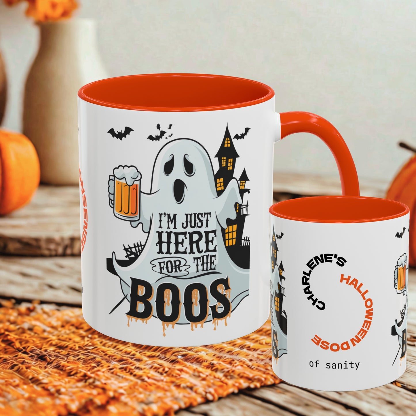 Two white ceramic mugs with orange interiors and handles, styled on a woven orange mat with autumn props. The larger mug features a Halloween graphic with a ghost, haunted house, bats, and the text 'I'M JUST HERE FOR THE BOOS'.