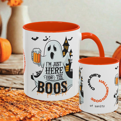 Two white ceramic mugs with orange interiors and handles, styled on a woven orange mat with autumn props. The larger mug features a Halloween graphic with a ghost, haunted house, bats, and the text 'I'M JUST HERE FOR THE BOOS'.