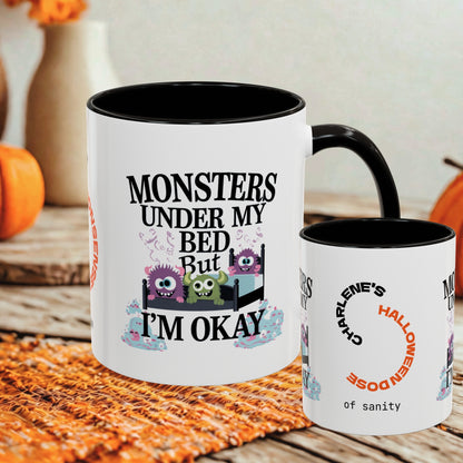 A white ceramic mug with a black interior and handle is shown on a rustic table, alongside a smaller side view of the same mug. The front mug features the text 'MONSTERS UNDER MY BED BUT I'M OKAY' above three cartoon monsters in a bed, and the side mug shows 'CHARLIE'S HALLOWEENDOSE of sanity' in curved orange and black text.