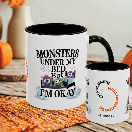 A white ceramic mug with a black interior and handle is shown on a rustic table, alongside a smaller side view of the same mug. The front mug features the text 'MONSTERS UNDER MY BED BUT I'M OKAY' above three cartoon monsters in a bed, and the side mug shows 'CHARLIE'S HALLOWEENDOSE of sanity' in curved orange and black text.