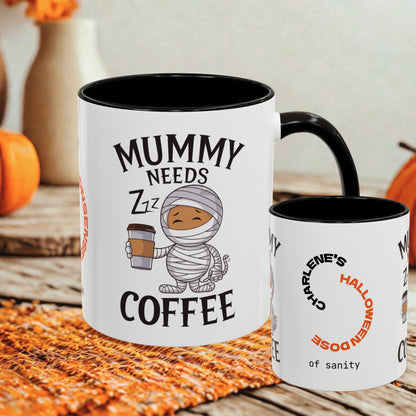Two white ceramic mugs with black interiors and handles, styled on a woven orange mat with autumn props in the background. The larger mug reads 'MUMMY NEEDS COFFEE' with a cartoon mummy holding a coffee cup, and the smaller mug reads 'CHARLENE'S HALLOWEEN DOSE of sanity'.