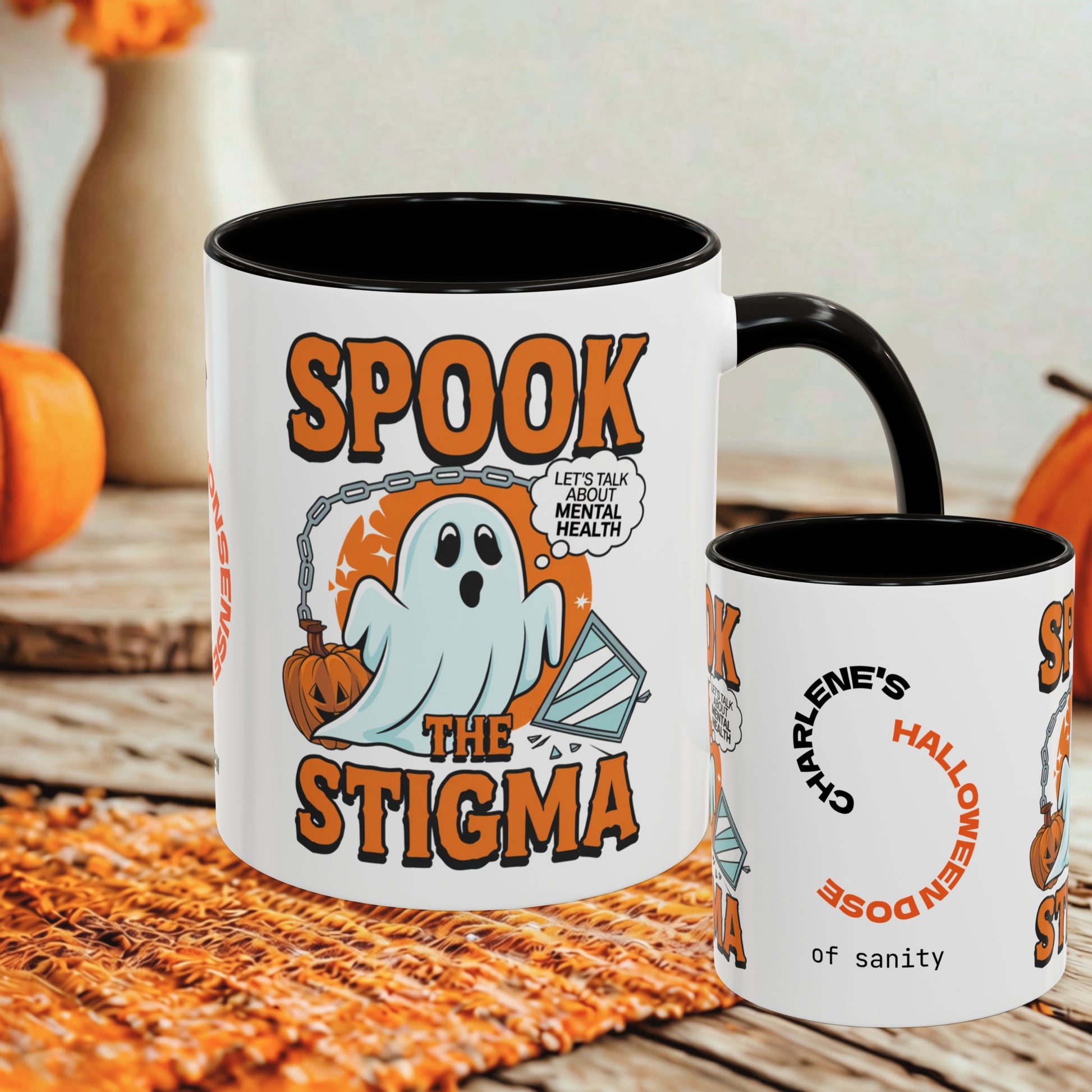 A white ceramic coffee mug with a black interior and handle, printed with a Halloween graphic reading 'SPOOK THE STIGMA' and a ghost holding an orange pumpkin. A smaller side view of the same mug is shown beside it on a rustic wooden surface with pumpkins and a woven mat in the background.