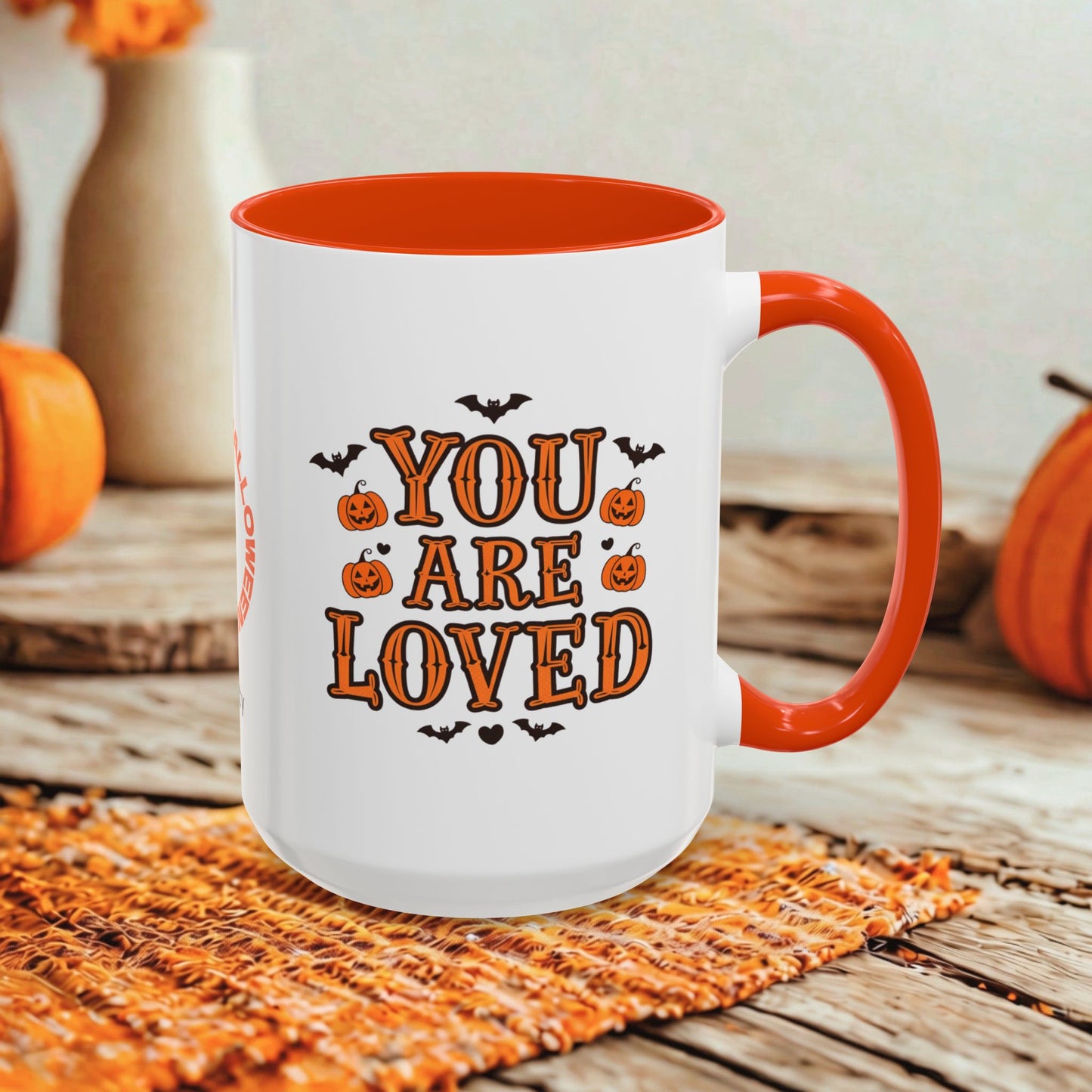 A white ceramic mug with an orange handle and interior, printed with the phrase 'YOU ARE LOVED' in orange lettering with small pumpkin and bat illustrations. The mug is styled on a woven orange placemat on a rustic wooden surface with seasonal pumpkins and vases in the background.