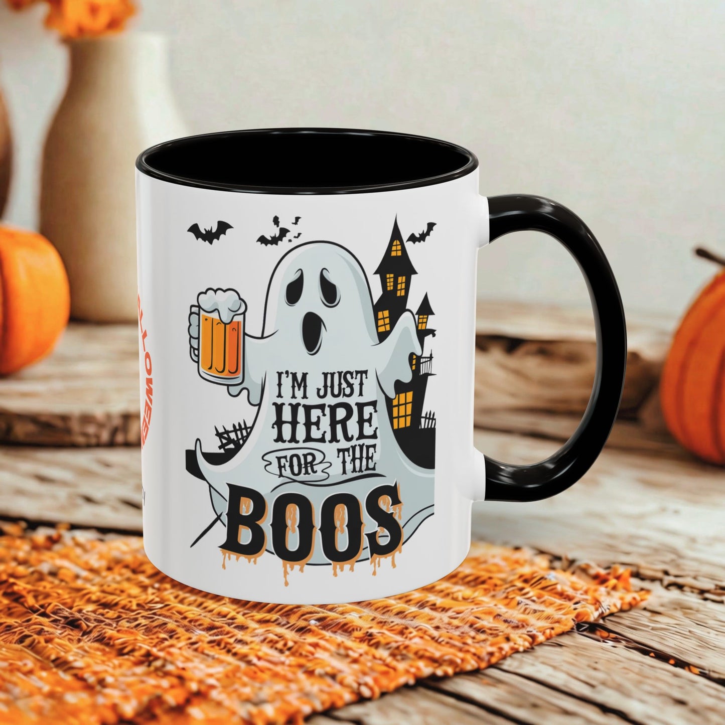 A white ceramic mug with a black handle and interior, printed with a Halloween graphic featuring a ghost, bats, a haunted house, and the text 'I'M JUST HERE FOR THE BOOS'. The mug is styled on a woven orange placemat with pumpkins and a rustic wooden table in the background.