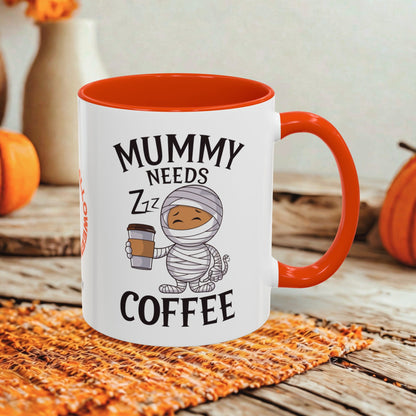 A white ceramic coffee mug with a bright orange handle and interior, printed with the phrase 'MUMMY NEEDS ZZZ COFFEE' and a cartoon mummy holding a coffee cup. The mug is styled on a woven orange placemat on a rustic wooden surface with pumpkins and a blurred background.