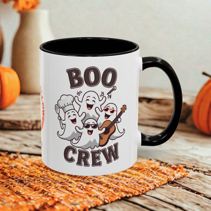 A white ceramic mug with a black handle and interior, printed with the words 'BOO CREW' and four cartoon ghosts in Halloween outfits, set on a rustic wooden surface with orange pumpkins and a woven mat in the background.