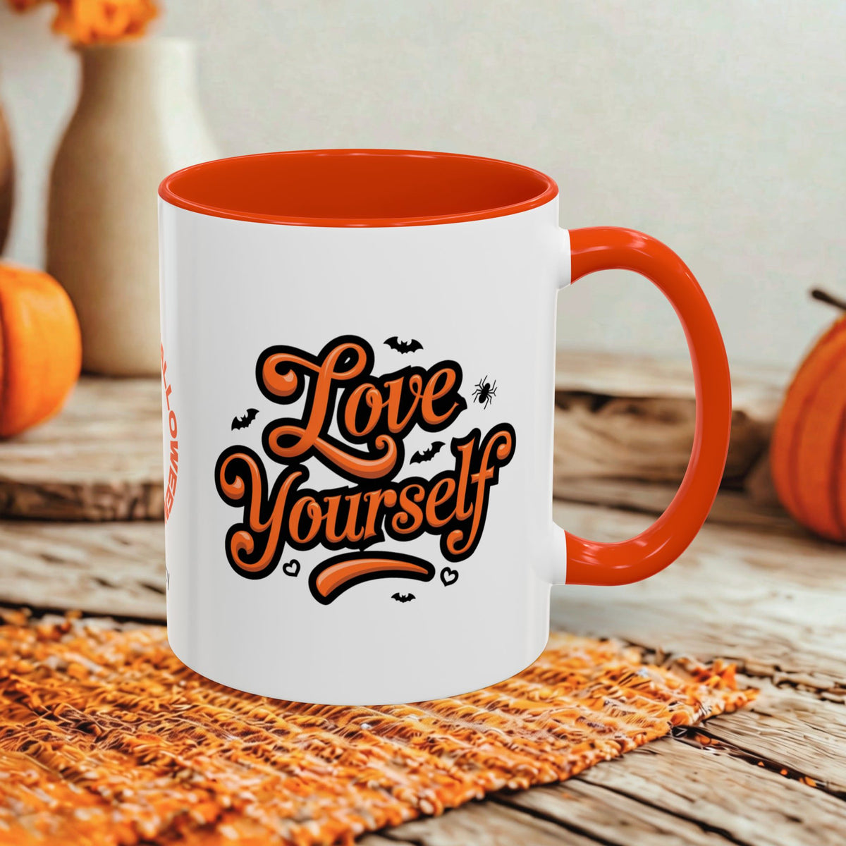 A white ceramic coffee mug with a bright orange handle and interior, featuring the phrase 'Love Yourself' in stylized orange script with small black bats and spider graphics. The mug is styled on a rustic wooden table with a woven placemat and orange pumpkins in the background.