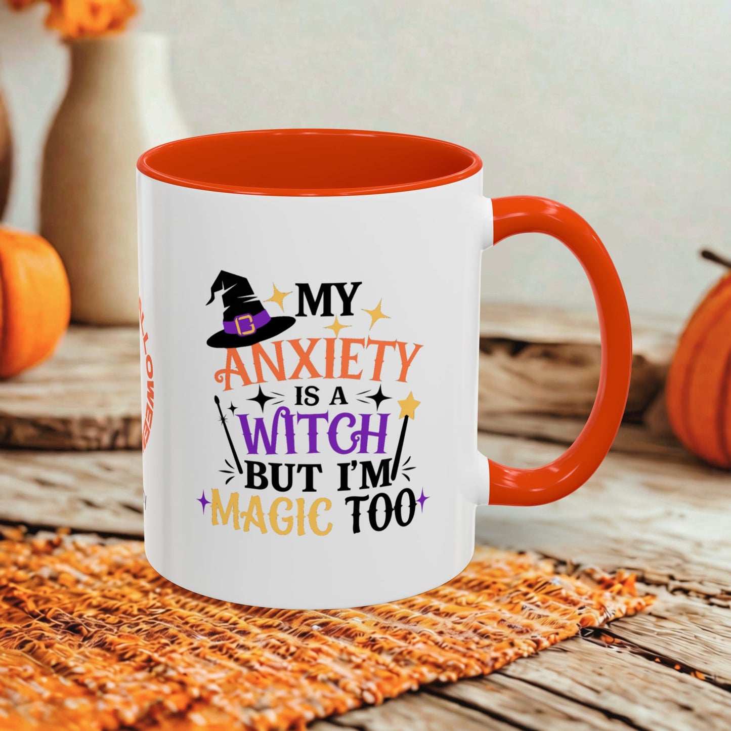 A white ceramic coffee mug with a bright orange handle and interior, featuring a Halloween-themed graphic that reads 'MY ANXIETY IS A WITCH BUT I'M MAGIC TOO' in orange, purple, and black lettering. The mug is styled on a rustic wooden surface with a woven placemat and blurred orange pumpkins in the background.
