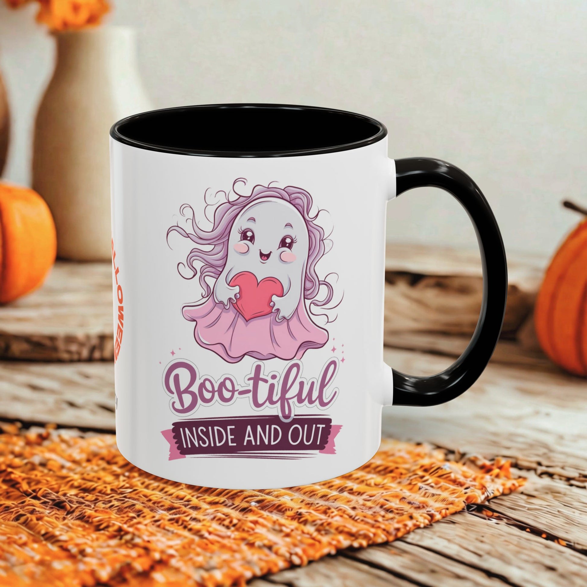 A white ceramic mug with a black handle and interior, featuring a cute pink ghost graphic and the text 'Boo-tiful INSIDE AND OUT.' The mug is styled on a woven orange placemat over a rustic wooden surface, with pumpkins and a soft neutral background visible.