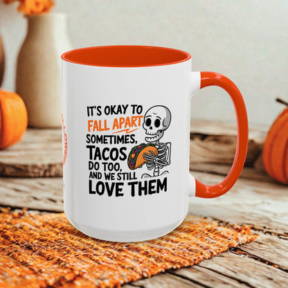 A white ceramic mug with a bright orange interior and handle, featuring a Halloween-themed printed design with a skeleton and the text 'IT'S OKAY TO FALL APART SOMETIMES, TACOS DO TOO, AND WE STILL LOVE THEM.' The mug is styled on a woven placemat over a rustic wooden surface with pumpkins and blurred decorative vases in the background.
