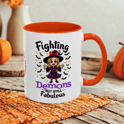 A white ceramic mug with a bright orange interior and handle, printed with a cartoon witch and the phrase 'Fighting Demons But Still Fabulous' in black and purple lettering. The mug is styled on a woven orange placemat with autumn pumpkins and a neutral wall in the background.