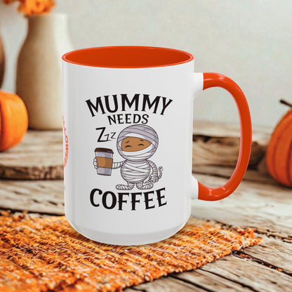 A white ceramic coffee mug with a bright orange handle and interior, printed with the text 'MUMMY NEEDS ZZZ COFFEE' and a cartoon mummy holding a coffee cup. The mug is styled on a woven orange placemat over a rustic wooden surface with pumpkins and blurred decorative vases in the background.