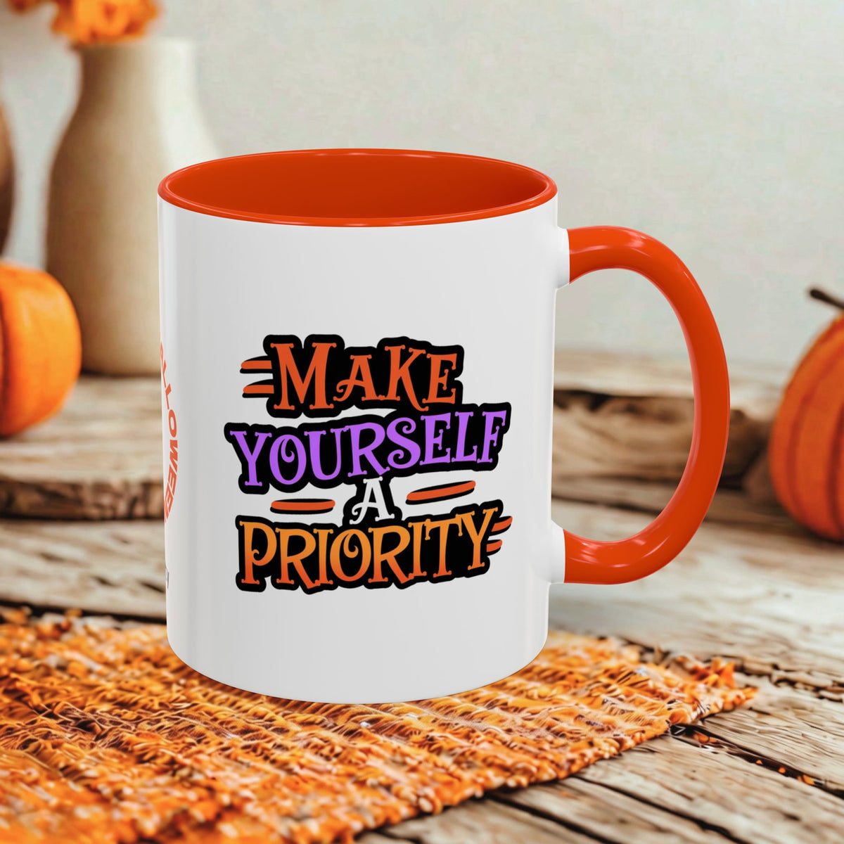 A white ceramic coffee mug with a bright orange handle and interior, featuring the printed text 'MAKE YOURSELF A PRIORITY' in stylized orange, purple, and black lettering. The mug is styled on a rustic wooden table with autumn-themed props like pumpkins and a woven placemat.