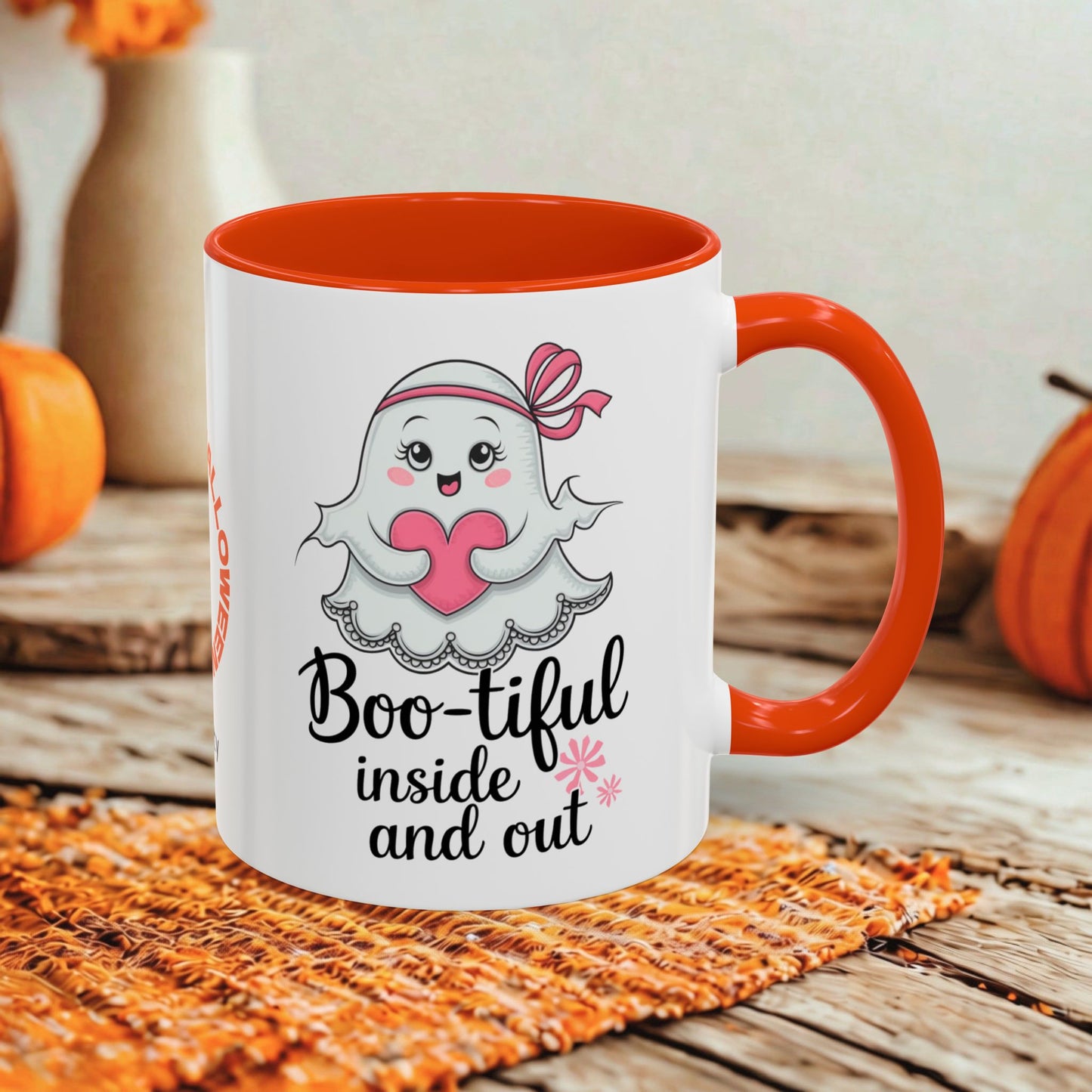 A white ceramic mug with a bright orange handle and interior, featuring a cute cartoon ghost holding a pink heart and the text 'Boo-tiful inside and out.' The mug is styled on a woven orange placemat over a rustic wooden surface, with pumpkins and a blurred vase in the background.