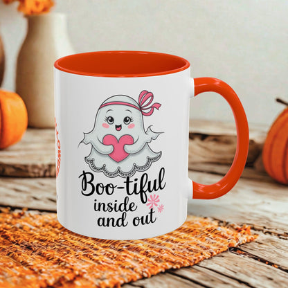 A white ceramic mug with a bright orange handle and interior, featuring a cute cartoon ghost holding a pink heart and the text 'Boo-tiful inside and out.' The mug is styled on a woven orange placemat over a rustic wooden surface, with pumpkins and a blurred vase in the background.