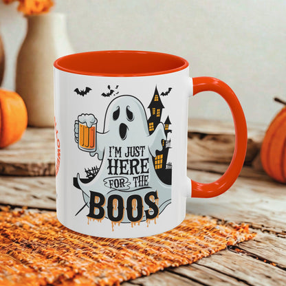 A white ceramic mug with a bright orange handle and interior, featuring a Halloween graphic with a ghost, haunted house, bats, and the text 'I'M JUST HERE FOR THE BOOS'. The mug is styled on a woven orange placemat on a rustic wooden table with pumpkins and a blurred background.