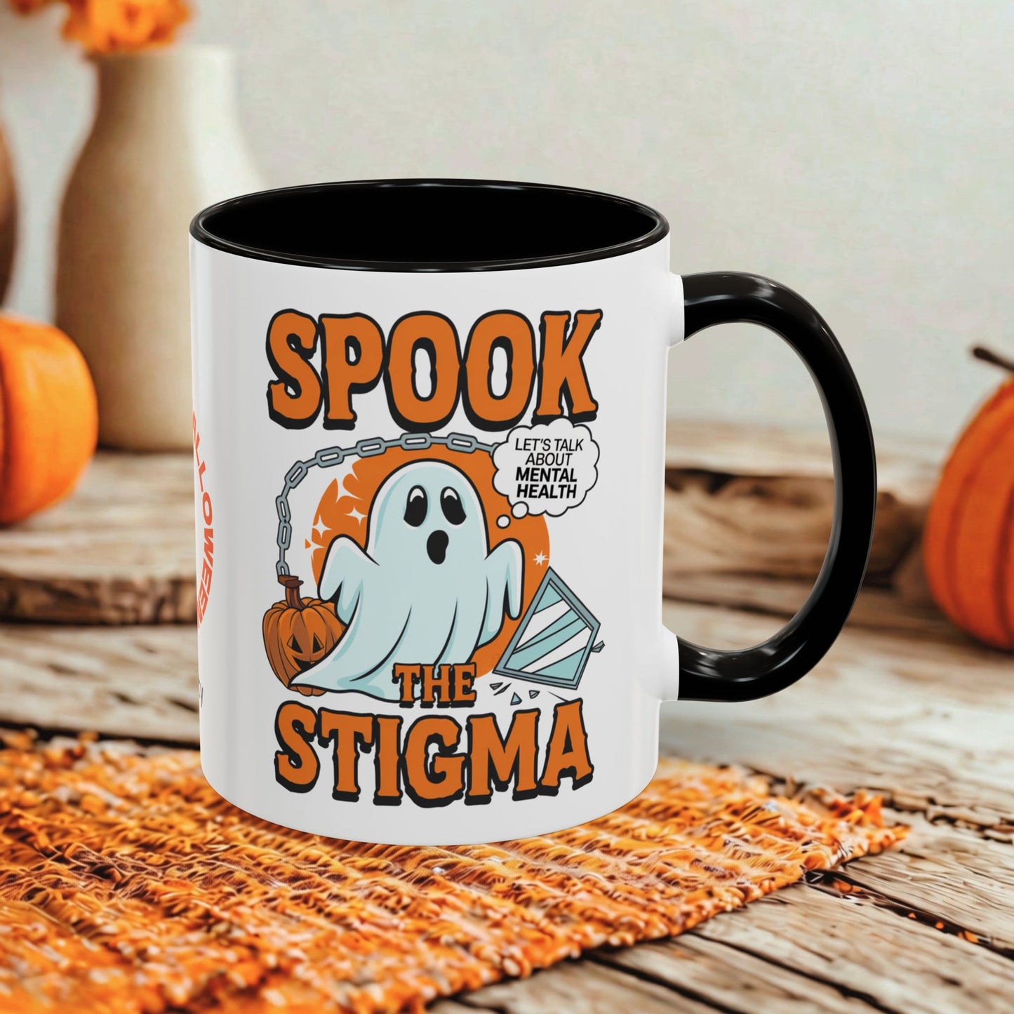 A white ceramic mug with a black handle and interior, printed with a Halloween graphic that reads 'SPOOK THE STIGMA' and shows a ghost, pumpkin, and a speech bubble. The mug is styled on a woven orange placemat on a rustic wooden table with blurred orange pumpkins and a neutral background.