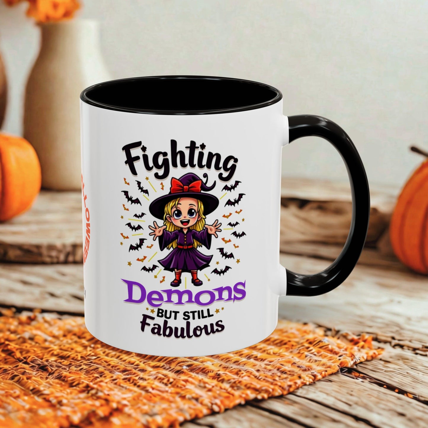A white ceramic mug with a black handle and interior, printed with a Halloween-themed graphic that reads 'Fighting Demons But Still Fabulous' around an illustration of a witch and bats. The mug is photographed on a rustic wooden surface with autumn props in the background.