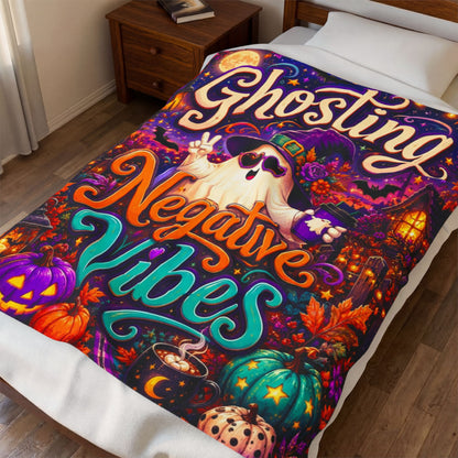 A rectangular Halloween-themed blanket displayed against a light wood plank background. The artwork features the words 'Ghosting Negative Vibes' in large decorative lettering, with a cartoon ghost wearing a witch hat and sunglasses, surrounded by pumpkins, a moonlit sky, a house, and lanterns.