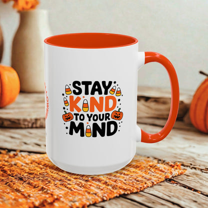 A white ceramic mug with a bright orange handle and interior, featuring a Halloween-themed graphic that reads 'STAY KIND TO YOUR MIND' with candy corn and jack-o'-lantern illustrations. The mug is styled on a rustic wooden surface with autumn props in the background.