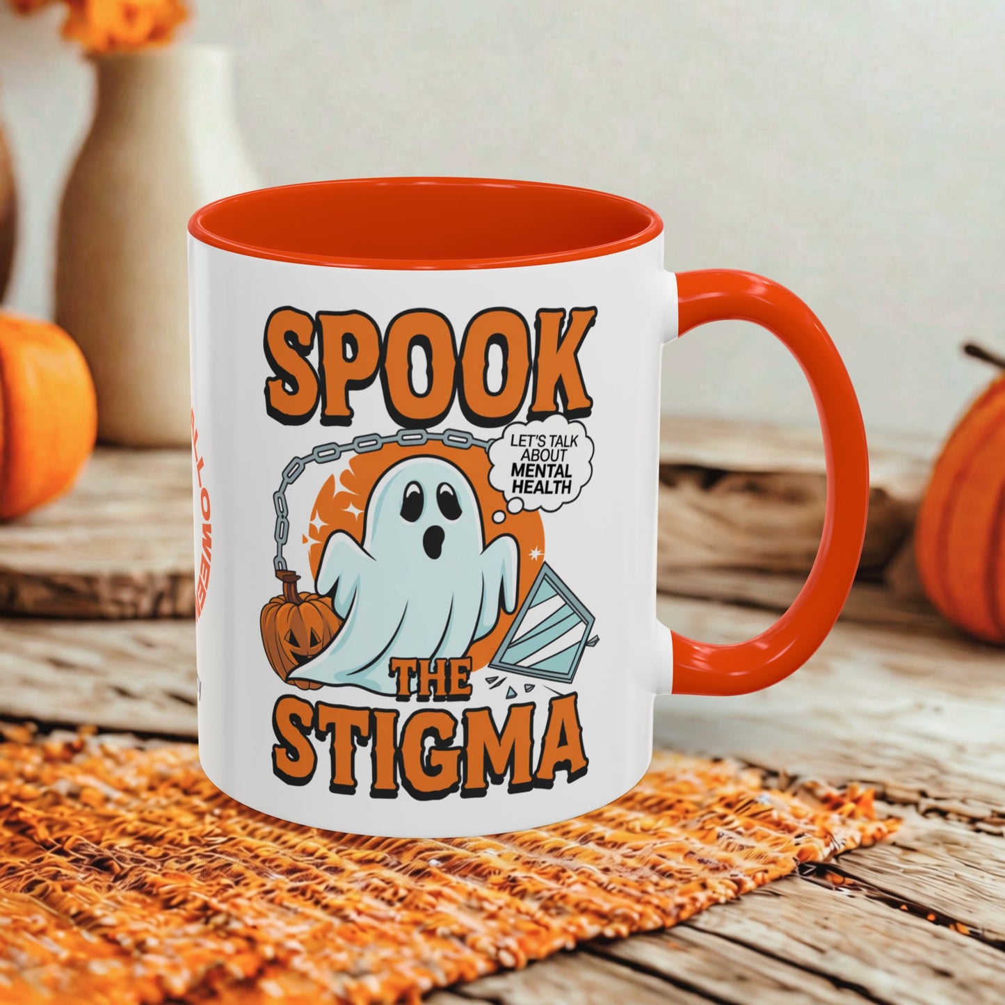 A white ceramic mug with a bright orange handle and interior, featuring a Halloween graphic that reads 'SPOOK THE STIGMA' with a ghost, pumpkin, and speech bubble. The mug is styled on a woven orange placemat over a rustic wooden surface with autumn props in the background.
