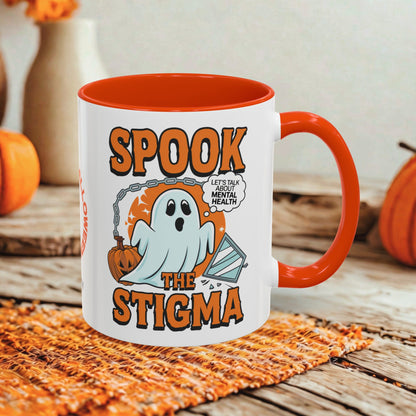 A white ceramic mug with a bright orange handle and interior, featuring a Halloween graphic that reads 'SPOOK THE STIGMA' with a ghost, pumpkin, and speech bubble. The mug is styled on a woven orange placemat over a rustic wooden surface with autumn props in the background.