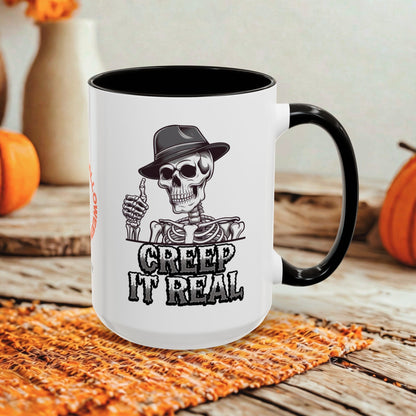 A white ceramic mug with a black handle and black interior, featuring a black-and-white graphic of a skeleton in a hat giving a thumbs-up with the text 'CREEP IT REAL'. The mug is photographed on a rustic wooden surface with autumn-themed props in the background.