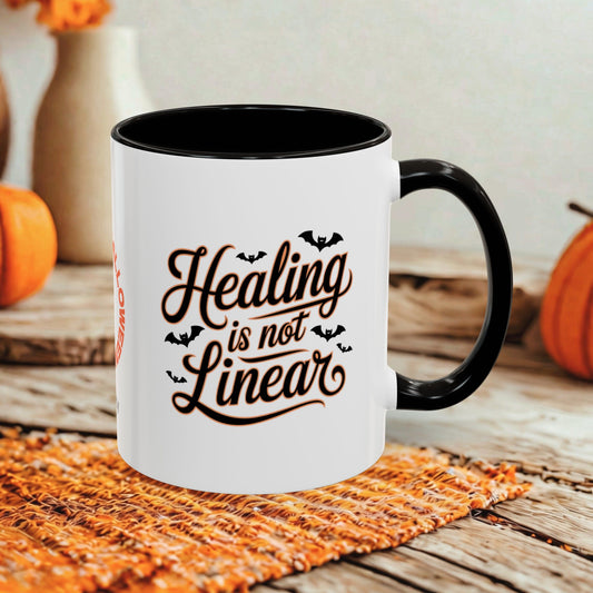 A white ceramic mug with a black handle and interior, printed with the phrase 'Healing is not Linear' in dark brown script with small bat silhouettes. The mug sits on a woven orange mat on a rustic wooden table, with pumpkins and a blurred indoor background.