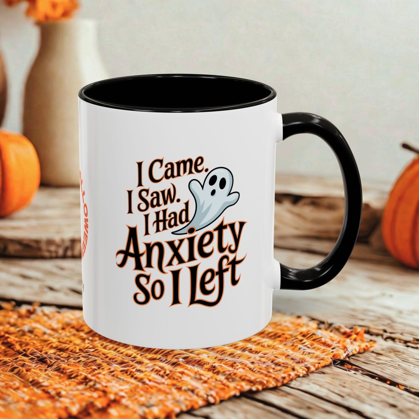 A white ceramic coffee mug with a black handle and interior, featuring a Halloween-themed graphic that reads 'I Came, I Saw, I Had Anxiety So I Left' alongside a small ghost illustration. The mug is styled on a woven orange placemat over a rustic wooden table, with pumpkins and a blurred background.