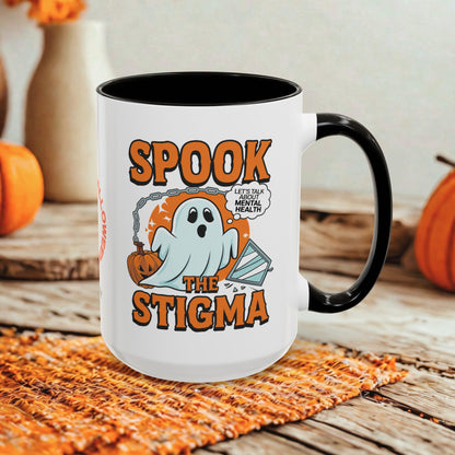 A white ceramic coffee mug with a black interior and handle, featuring a Halloween graphic that reads 'SPOOK THE STIGMA' with a ghost, pumpkin, and speech bubble saying 'LET'STALK ABOUT MENTAL HEALTH'. The mug is styled on a woven orange placemat over a rustic wooden surface with pumpkins and blurred background decor.