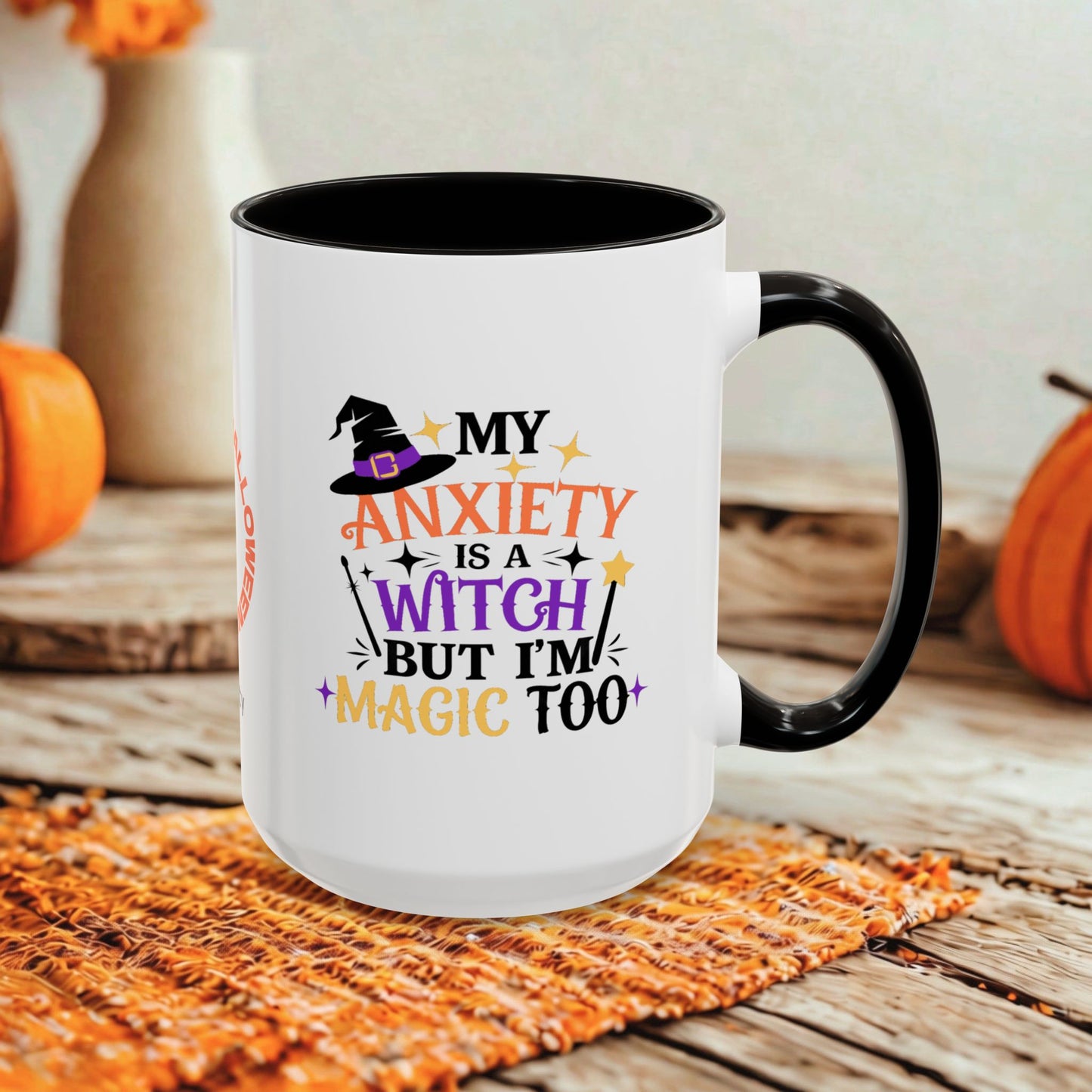 A white ceramic mug with a black handle and black interior, printed with the phrase 'MY ANXIETY IS A WITCH BUT I'M MAGIC TOO' in orange, purple, and black lettering with a witch hat graphic. The mug is styled on a rustic wooden surface with autumn props including pumpkins and a woven placemat.