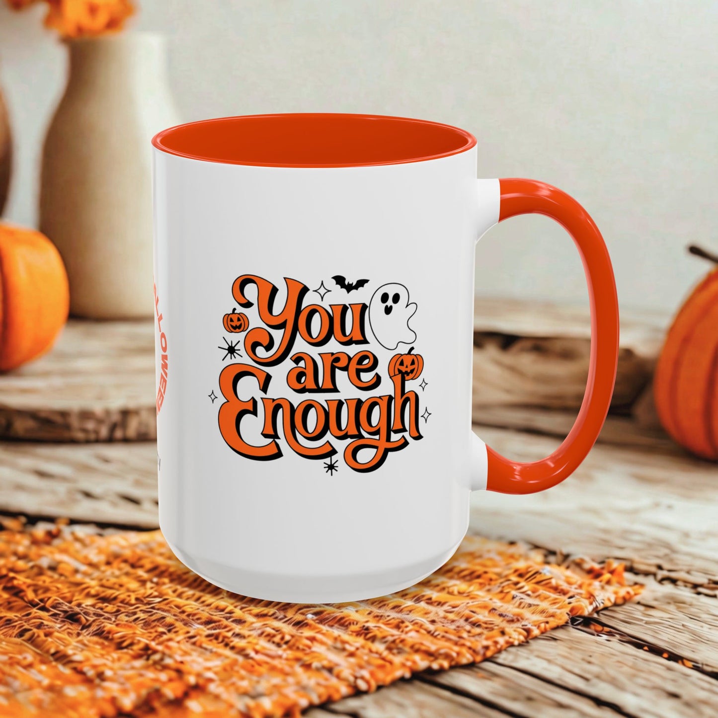 A white ceramic mug with a bright orange handle and interior, featuring the phrase 'You are Enough' in orange script with pumpkins, a ghost, and bats. The mug is styled on a woven orange placemat on a rustic wooden table with orange pumpkins and neutral background decor.