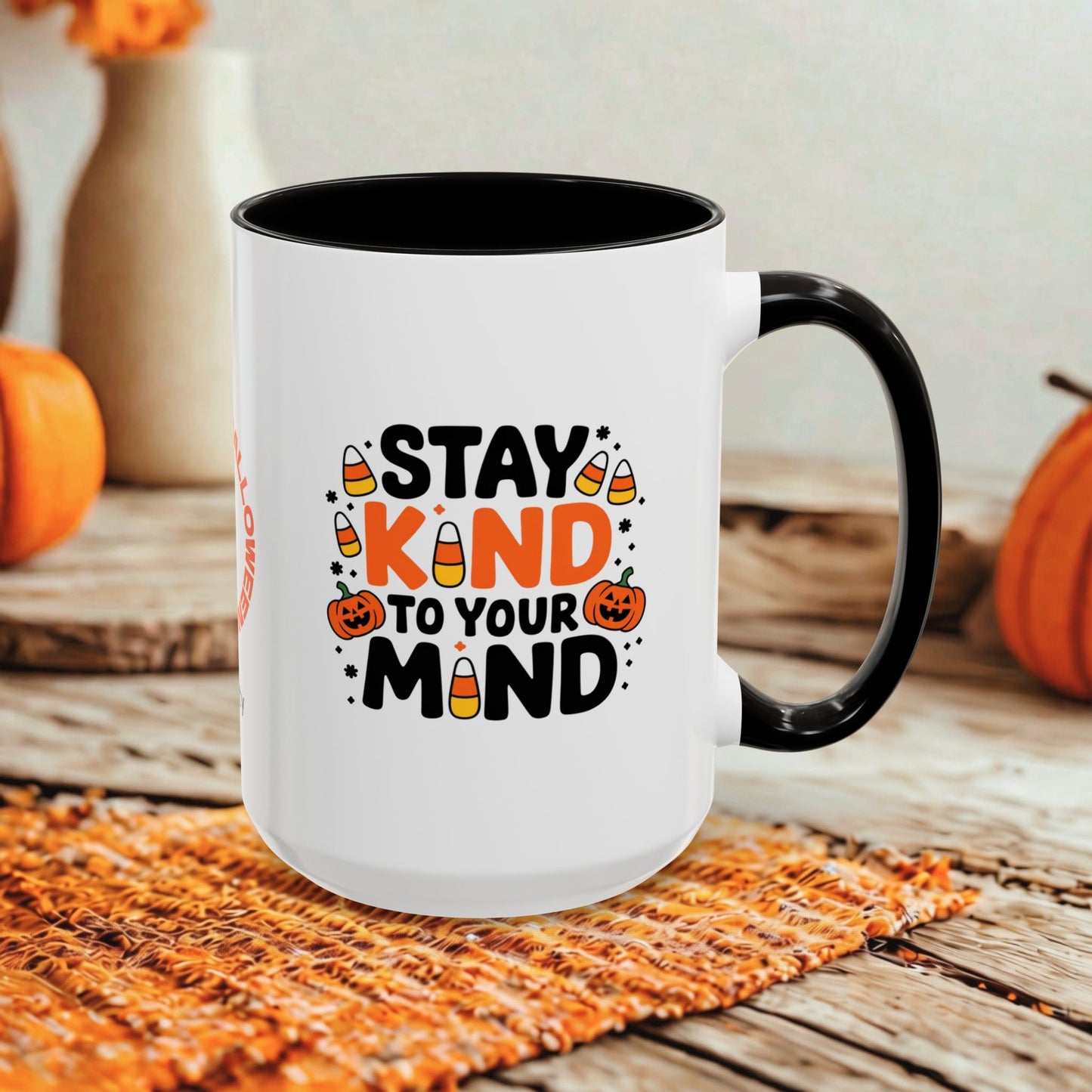 A white ceramic mug with a black handle and black interior, printed with the phrase 'STAY KIND TO YOUR MIND' in black and orange lettering with candy corn and jack-o'-lantern illustrations. The mug is set on a woven orange placemat on a rustic wooden table, with blurred orange pumpkins and neutral background decor.