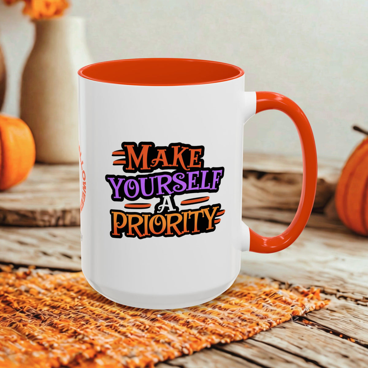 A white ceramic coffee mug with a bright orange handle and interior, featuring the printed text 'MAKE YOURSELF A PRIORITY' in orange, purple, and black. The mug is styled on a woven orange placemat on a rustic wooden table with autumn-themed background props.