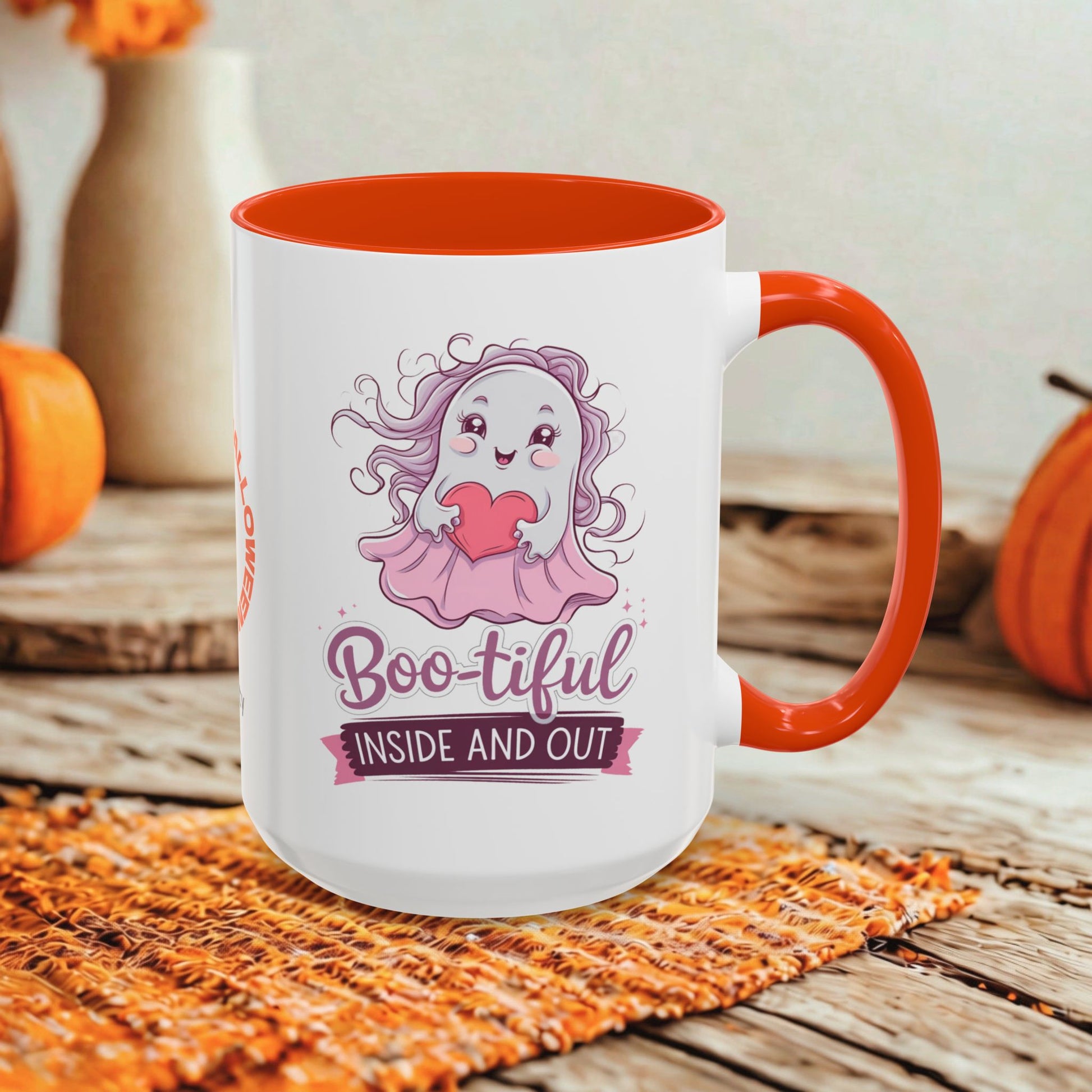 A white ceramic mug with a bright orange handle and interior, featuring a cute cartoon ghost graphic and the text 'Boo-tiful INSIDE AND OUT.' The mug is styled on a rustic wooden surface with autumn props including pumpkins and a woven placemat.
