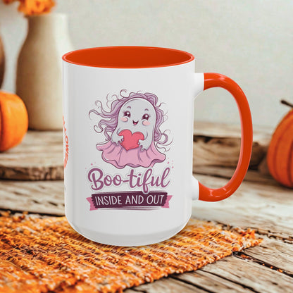 A white ceramic mug with a bright orange handle and interior, featuring a cute cartoon ghost graphic and the text 'Boo-tiful INSIDE AND OUT.' The mug is styled on a rustic wooden surface with autumn props including pumpkins and a woven placemat.