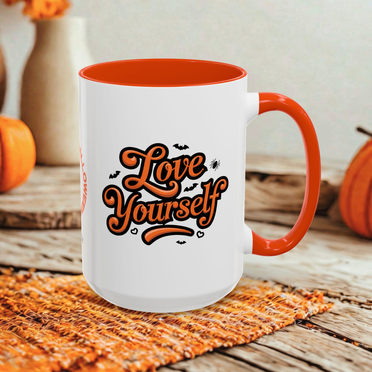 A white ceramic mug with an orange interior, handle, and rim, featuring the phrase 'Love Yourself' in orange script with small bat and spider graphics. The mug is styled on a woven placemat on a rustic wooden table with orange pumpkins and blurred decorative vases in the background.
