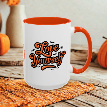 A white ceramic mug with an orange interior, handle, and rim, featuring the phrase 'Love Yourself' in orange script with small bat and spider graphics. The mug is styled on a woven placemat on a rustic wooden table with orange pumpkins and blurred decorative vases in the background.