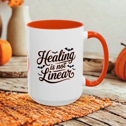 A white ceramic mug with a bright orange handle and interior, printed with the phrase 'Healing is not Linear' in dark brown script with small bat silhouettes. The mug sits on a woven orange placemat on a rustic wooden surface, with pumpkins and a blurred indoor background.