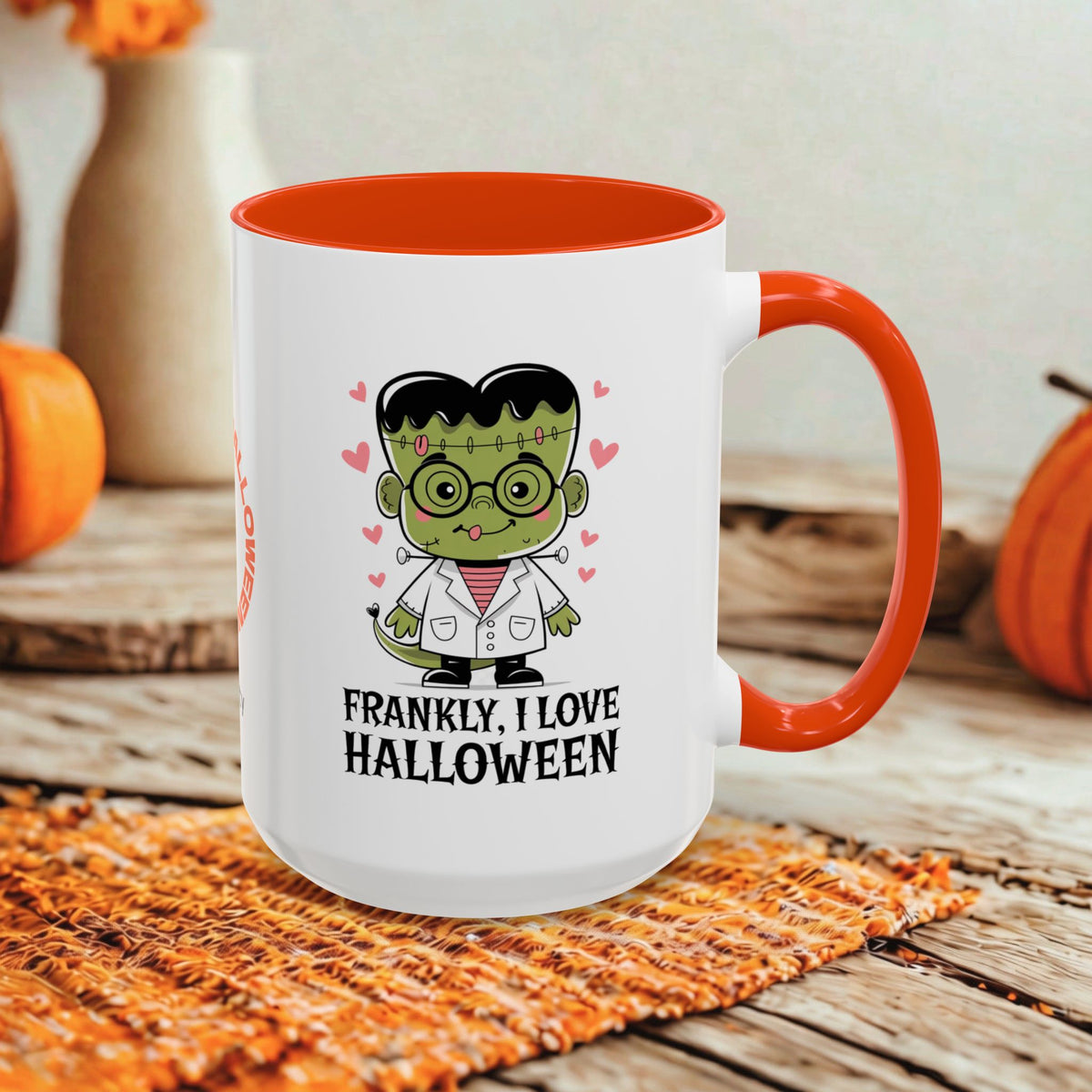A white ceramic mug with a bright orange handle and interior, featuring a cartoon Frankenstein character and the text 'FRANKLY, I LOVE HALLOWEEN' on the front. The mug is styled on a rustic wooden surface with autumn-themed props including pumpkins and a woven placemat.