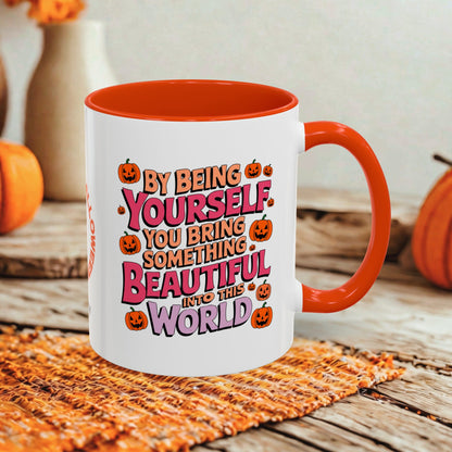 A white ceramic coffee mug with a bright orange handle and interior, featuring a colorful Halloween-themed graphic that reads 'BY BEING YOURSELF YOU BRING SOMETHING BEAUTIFUL INTO THE WORLD' surrounded by pumpkins. The mug is styled on a rustic wooden table with a woven placemat and blurred orange pumpkins in the background.